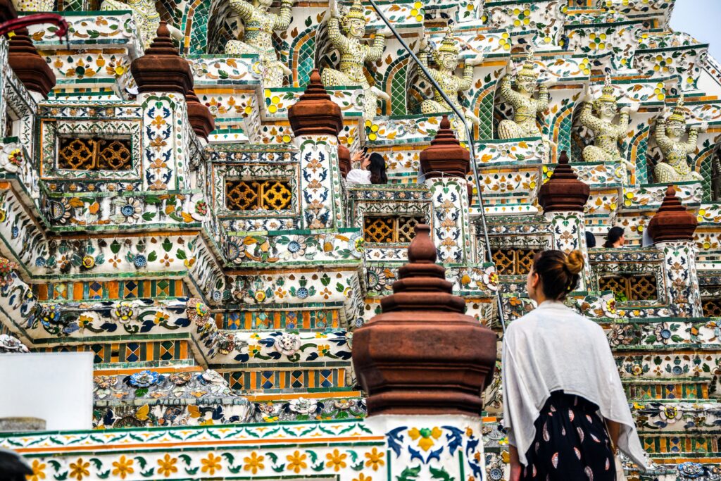 Détail coloré des prangs du Wat Arun à Bangkok, décorés de porcelaines et statues mythologiques, avec une visiteuse observant les ornements.